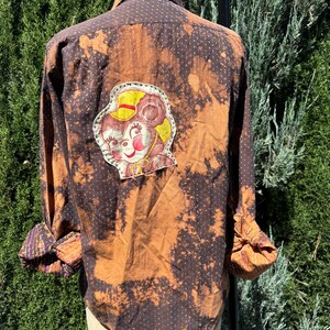 Puede incluir: Una camisa de manga larga marr&oacute;n y negra con un patr&oacute;n de lunares blancos. La camisa tiene un dise&ntilde;o salpicado de lej&iacute;a y un parche de rat&oacute;n de dibujos animados en la espalda.