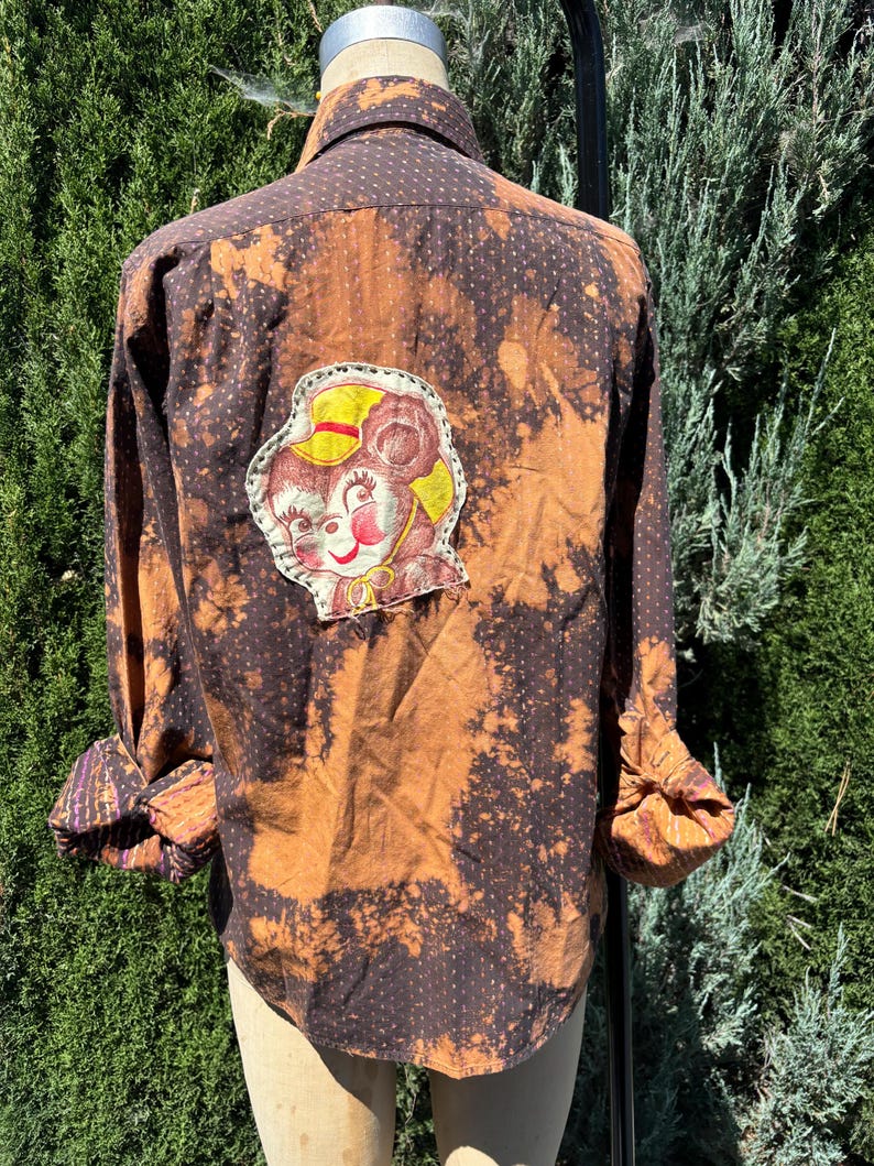 Puede incluir: Una camisa de manga larga marr&oacute;n y negra con un patr&oacute;n de lunares blancos. La camisa tiene un dise&ntilde;o salpicado de lej&iacute;a y un parche de rat&oacute;n de dibujos animados en la espalda.