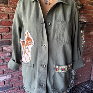 May include: A green utility jacket with a deer patch on the left side and a floral embroidered trim on the right pocket. The jacket has a button closure and a collar.