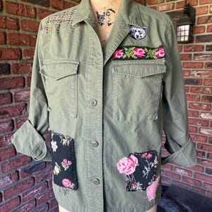 May include: A green utility jacket with various patches and embroidery. The patches include floral designs, a black and white animal, and a black and white floral design. The jacket has a button closure and two flap pockets.