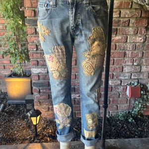 May include: Light blue denim jeans with decorative patches in various floral and patterned fabrics. The jeans have a classic five-pocket design, a button and zipper closure, and are cuffed at the ankles. The patches are in shades of beige, pink, and green.