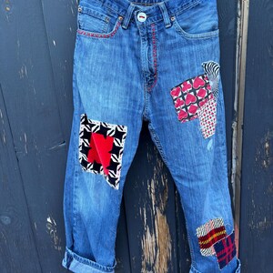May include: Blue denim jeans with decorative patches in various patterns and colors. Patches include red hearts, black and white geometric shapes, and plaid. The jeans have a red stitched detail on the pockets and cuffs.
