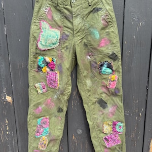 May include: Olive green pants with colorful patches and paint splatters. The patches are various shapes and sizes, with floral and abstract designs. The pants have a button closure and pockets, and are hanging on a wooden hanger.