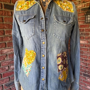 May include: A light blue denim shirt with a floral pattern on the collar and pockets. The shirt has a button-up front and long sleeves.