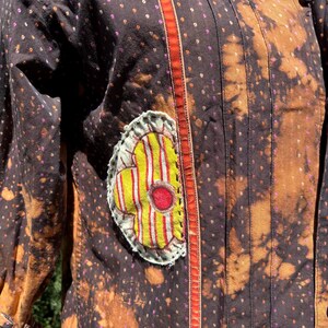 Puede incluir: Una camisa de estampado marr&oacute;n y naranja con una aplicaci&oacute;n de flor amarilla y roja. La camisa tiene un ribete rojo y naranja en la parte delantera.