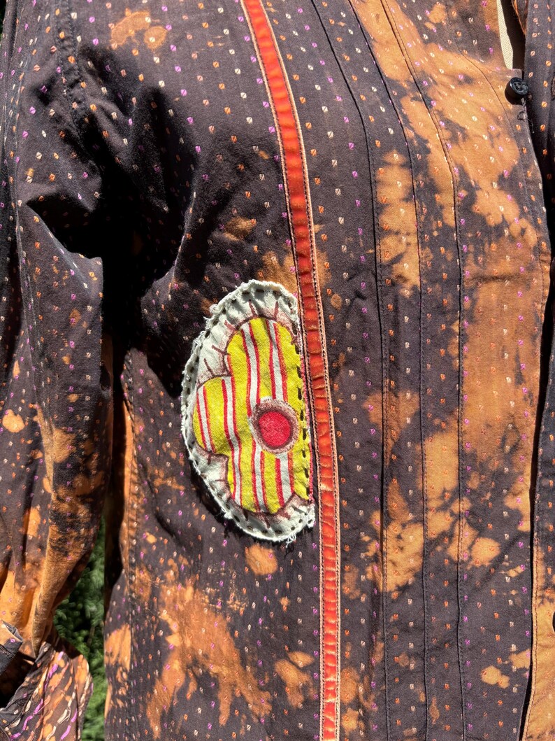 Puede incluir: Una camisa de estampado marr&oacute;n y naranja con una aplicaci&oacute;n de flor amarilla y roja. La camisa tiene un ribete rojo y naranja en la parte delantera.