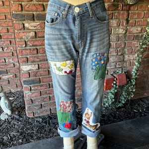 May include: Blue denim jeans with patchwork details. The patches are made of different fabrics and feature various designs, including a floral pattern, a bird design, and a heart design.