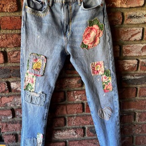 May include: Light blue denim jeans with floral patches and a heart-shaped patch. The jeans have a classic five-pocket design and a straight leg fit. The patches feature pink and red roses and other floral patterns.