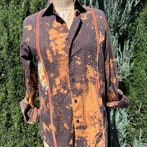 Puede incluir: Una camisa de botones de manga larga de color marr&oacute;n y naranja con un patr&oacute;n de lunares blancos y rayas rojas y blancas en la parte delantera. La camisa tiene un aspecto descolorido, blanqueado y un peque&ntilde;o parche bordado en el lado izquierdo.