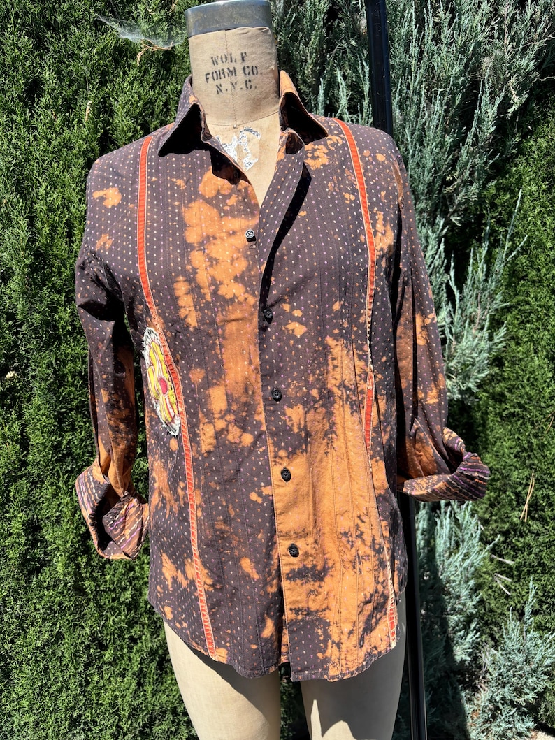 Puede incluir: Una camisa de botones de manga larga de color marr&oacute;n y naranja con un patr&oacute;n de lunares blancos y rayas rojas y blancas en la parte delantera. La camisa tiene un aspecto descolorido, blanqueado y un peque&ntilde;o parche bordado en el lado izquierdo.