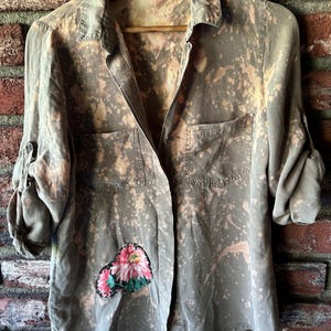 May include: A green button-up shirt with a floral patch on the left side. The shirt has a bleach-splattered design.