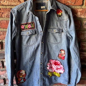 May include: Light blue denim shirt adorned with colorful embroidered patches. The patches feature floral designs and a teddy bear. The shirt has two chest pockets and a button-down front. The shirt is hanging on a wooden hanger against a brick wall.