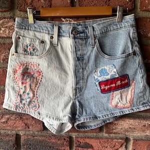 May include: Light and medium wash denim shorts with patchwork details. The shorts feature floral and patterned fabric patches, and a red patch that reads "Laguna Beach". The shorts are hanging on a wooden hanger against a brick wall.