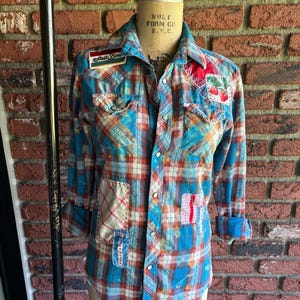 May include: A button-down plaid shirt in shades of blue, red, and orange, displayed on a mannequin. The shirt features patches and embellishments, including a patch that says "Stella Dore" and a cherry design. The sleeves are rolled up.