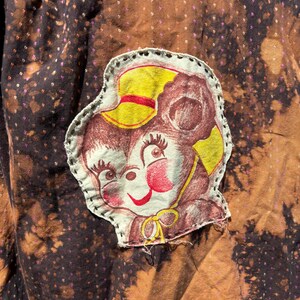 Puede incluir: Una tela marr&oacute;n con un parche de oso de dibujos animados blanco y marr&oacute;n. El oso lleva un sombrero amarillo con una banda roja. La tela tiene un patr&oacute;n moteado marr&oacute;n y negro.