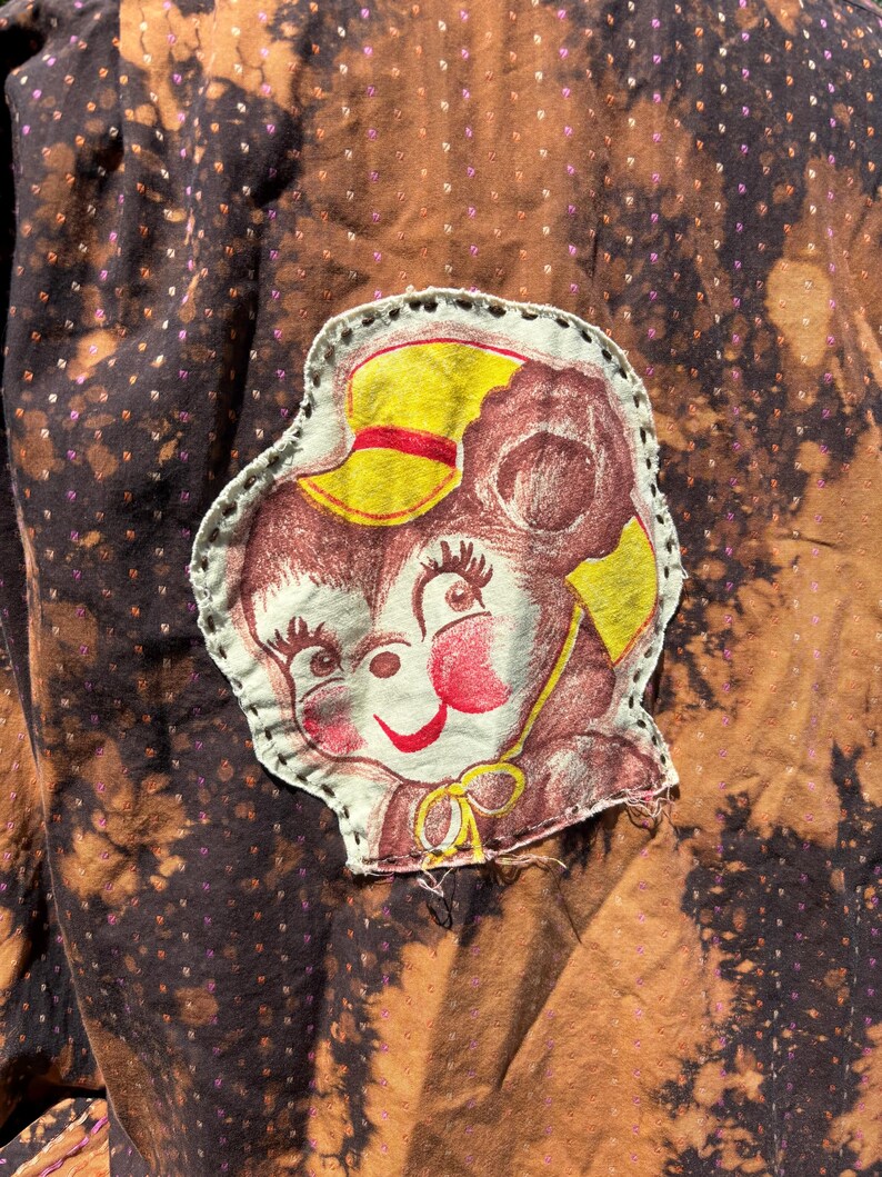 Puede incluir: Una tela marr&oacute;n con un parche de oso de dibujos animados blanco y marr&oacute;n. El oso lleva un sombrero amarillo con una banda roja. La tela tiene un patr&oacute;n moteado marr&oacute;n y negro.