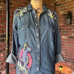 May include: A denim shirt with long sleeves, featuring a colorful embroidered design of a bird on the front and floral accents on the shoulders. The shirt has a button-down closure and two chest pockets.