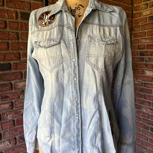 May include: A light blue denim shirt with a space shuttle patch on the left chest and a black and white graphic on the right sleeve. The shirt has a button-down front and long sleeves.