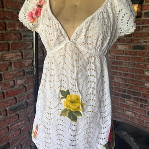 May include: White eyelet dress with a V-neckline and short sleeves. The dress features floral embroidery in pink, red, and yellow. The waist is cinched with an elastic band. The dress is displayed on a mannequin.