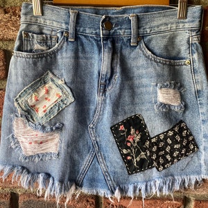 May include: A light blue denim mini skirt with frayed edges and various fabric patches sewn on. The patches feature floral and geometric patterns in shades of pink, white, and black.