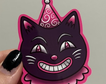 Puede incluir: Una pegatina con un gato de dibujos animados sonriente con un gorro de fiesta. El gato es morado con detalles rosas, incluyendo el gorro, los ojos, la nariz y el borde festoneado. Los bigotes y dientes blancos completan el diseño.