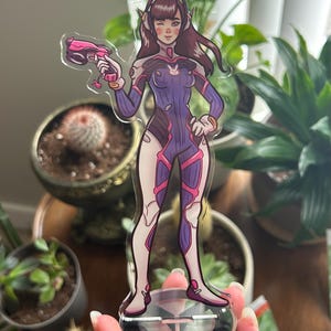 May include: A clear acrylic standee featuring a cartoon character with brown hair, wearing a purple and white bodysuit with pink accents, holding a pink blaster. The figure is mounted on a circular base with a pink and black geometric design.