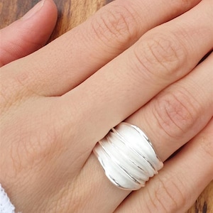 Könnte beinhalten: Ein silberner Ring mit einem strukturierten, wellenartigen Design. Der Ring wird an einem Finger getragen und zeigt seine glatte, reflektierende Oberfläche. Das Design des Rings weist mehrere parallele Rippen auf, die eine einzigartige Ästhetik erzeugen. Der Ring ist ein Schmuckstück.