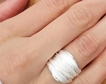 May include: A silver ring with a textured, wave-like design. The ring is worn on a person's finger, showcasing its smooth, reflective surface. The ring's design features multiple parallel ridges, creating a unique and eye-catching aesthetic. The ring is a piece of jewelry.