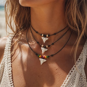 Op de afbeelding: Een set van drie gelaagde kettingen met witte haaientandhangers aan dunne zwarte koorden. De bovenste ketting heeft zwarte en beige kralen, de middelste heeft bruine kralen en de onderste ketting heeft rode, gele en groene accentkralen.