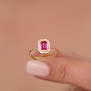 May include: A gold ring featuring a red emerald-cut gemstone with a halo of smaller diamonds.
