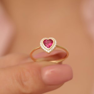 May include: A gold ring featuring a heart-shaped, ruby-colored gemstone, surrounded by a halo of small, clear gemstones. The ring is held up against a soft, pink background.