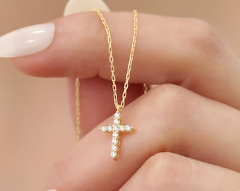 Op de afbeelding: Een goudkleurige ketting met een kruisvormige hanger. Het kruis is versierd met kleine, heldere edelstenen. De ketting wordt omhoog gehouden, waarbij de fijne ketting en de sprankelende details van het kruis te zien zijn. Een religieus symbool.