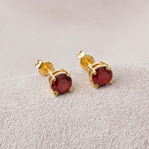 May include: A pair of gold stud earrings with red gemstone accents. The earrings are displayed on a light beige fabric surface.