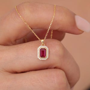 Könnte beinhalten: Eine goldene Kettenkette mit einem roten Edelstein-Anhänger. Der Anhänger ist von einem Heiligenschein aus kleineren, klaren Edelsteinen umgeben.