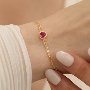 May include: A delicate gold chain bracelet featuring a teardrop-shaped ruby gemstone surrounded by smaller diamonds.