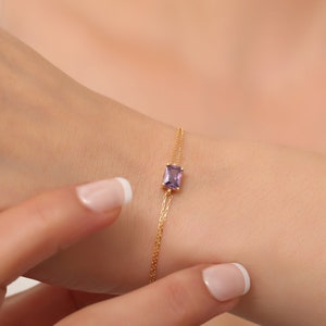 May include: A delicate gold chain bracelet with a single purple gemstone in a rectangular shape.