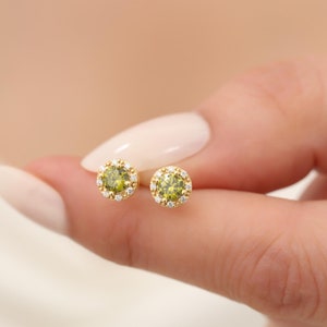 Op de afbeelding: Een paar gouden oorbellen met een groene edelsteen in het midden en een halo van kleinere witte edelstenen.