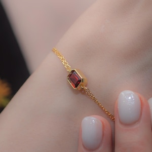Op de afbeelding: Een delicate gouden armband met een rechthoekige, dieprode edelsteen. De ketting van de armband is fijn en dubbelstrengs, met de edelsteen gezet in een gouden bezel. De armband wordt getoond op een pols met verzorgde nagels.