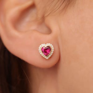 Könnte beinhalten: Ein Paar goldene herzförmige Ohrringe mit einem rosafarbenen Edelstein in der Mitte und einem Heiligenschein aus kleinen weißen Edelsteinen am Rand.