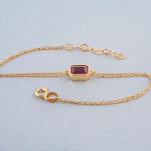 Op de afbeelding: Een gouden kettingarmband met een enkele rode edelsteen in een gouden frame.