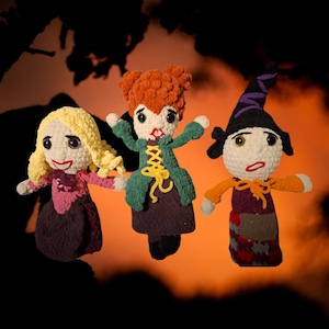 Może przedstawiać: Trzy ręcznie robione lalki na szydełku przedstawiające postacie z filmu Halloween. Lalki mają włosy z przędzy w kolorze blond, czerwonym i czarnym. Każda lalka nosi inny strój w odcieniach fioletu, zieleni i pomarańczy. Tło jest pomarańczowe i czarne.