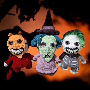 Pode incluir: Três bonecas de crochê feitas à mão com características assustadoras e exageradas. Uma é laranja e vermelha com um sorriso diabólico, outra é verde com cabelo azul e um chapéu de bruxa, e a terceira é branca e cinza com cabelo verde e um design esquelético.