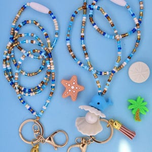 May include: Two beaded lanyards featuring blue, white, and metallic gold seed beads with gold-tone lobster clasps. One lanyard includes a blue whale charm, a white shell with a pearl, and a pink tassel. The other features a blue tassel.