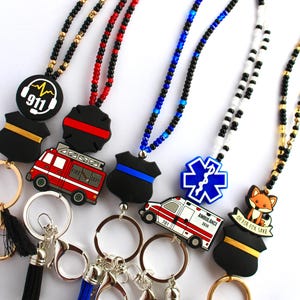 May include: A collection of beaded lanyards and keychains with emergency service-themed charms. Charms include a 911 logo, fire truck, ambulance, and police badge. Keychains have tassels in various colors.