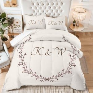 May include: A white comforter set with embroidered brown initials "K & W" and a matching floral wreath design. Two matching pillowcases also feature the initials. The bed is set against a tufted headboard.