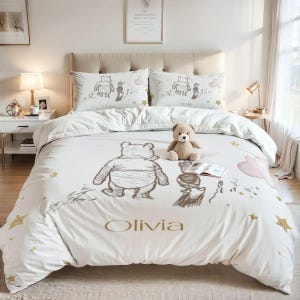Puede incluir: Juego de cama blanco con una ilustración de oso de dibujos animados y el nombre "Olivia" en letras doradas. El juego incluye una funda nórdica y dos fundas de almohada. Un oso de peluche y un libro abierto están en la cama. Estrellas doradas y nubes rosas también forman parte del diseño.