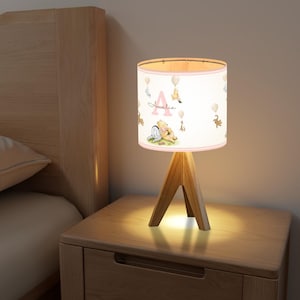 May include: A pink and white Winnie the Pooh themed lampshade with the name Amelia printed on it. The lampshade is on a wooden tripod base.