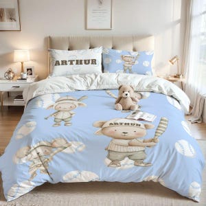 May include: A light blue bedding set featuring a repeating pattern of cartoon bears dressed in baseball uniforms holding bats and baseballs. The pillowcase displays the name ARTHUR in bold letters. A plush teddy bear sits on the center of the bed.