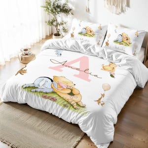 May include: White bedding set with a pink letter A and the name Amelia. The bedding features a Winnie the Pooh and Piglet illustration with a yellow balloon and other Winnie the Pooh characters.