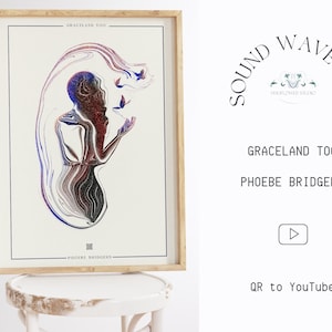 May include: A framed print of a digital art piece featuring a person with a swirling, abstract design in shades of blue, red, and black. The artwork is titled "Graceland Too" by Phoebe Bridgers. The print is on a white background and is framed in a light wood frame.